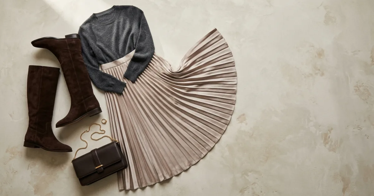 Champagne satin accordion pleated midi skirt in a gentle fan arc with charcoal cashmere crewneck top, chocolate suede tall boots, and leather clutch on a champagne limewash plaster surface